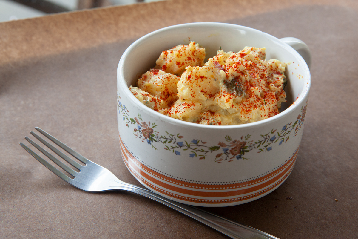 Bowl of potato salad garnished with paprika, served in a floral mug with a fork beside it on brown paper